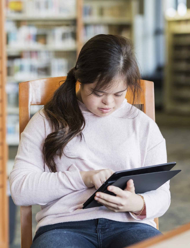 A young person sitting in a library and looking down at a tablet
